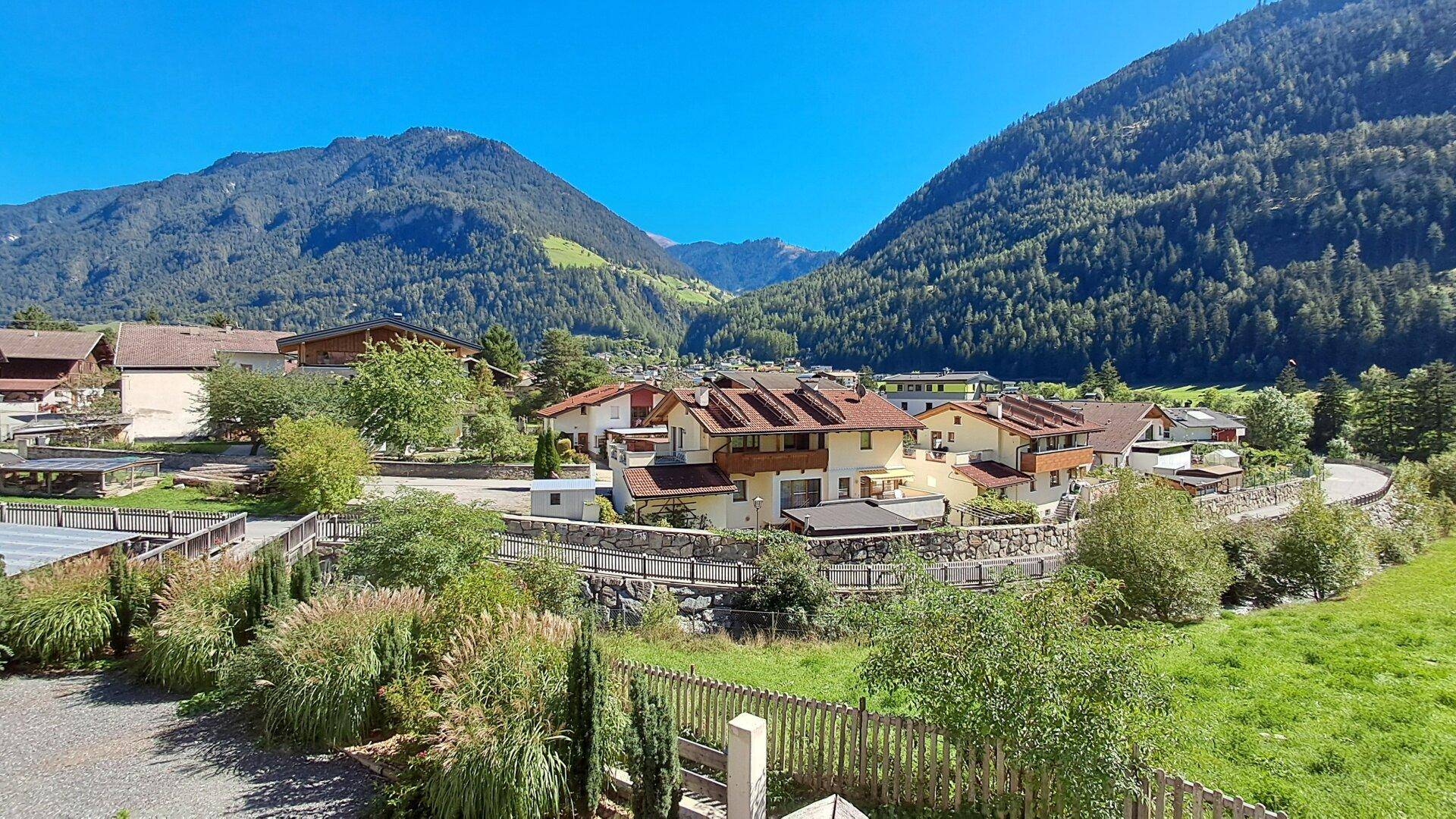 Breiter Panoramablick auf ein Dorf in einer alpinen Landschaft mit grünen Wiesen und hohen Bergen.