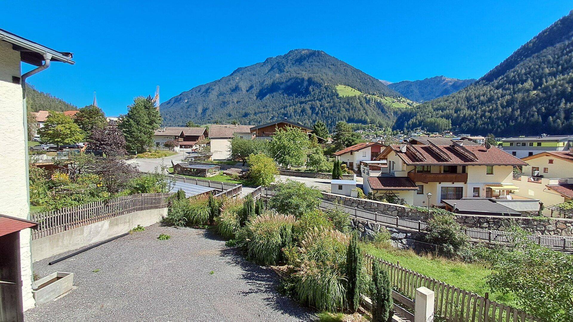 Blick auf ein Dorf mit grünen Landschaften und Bergen, teilweise verdeckt von einem Gebäude auf der linken Seite.