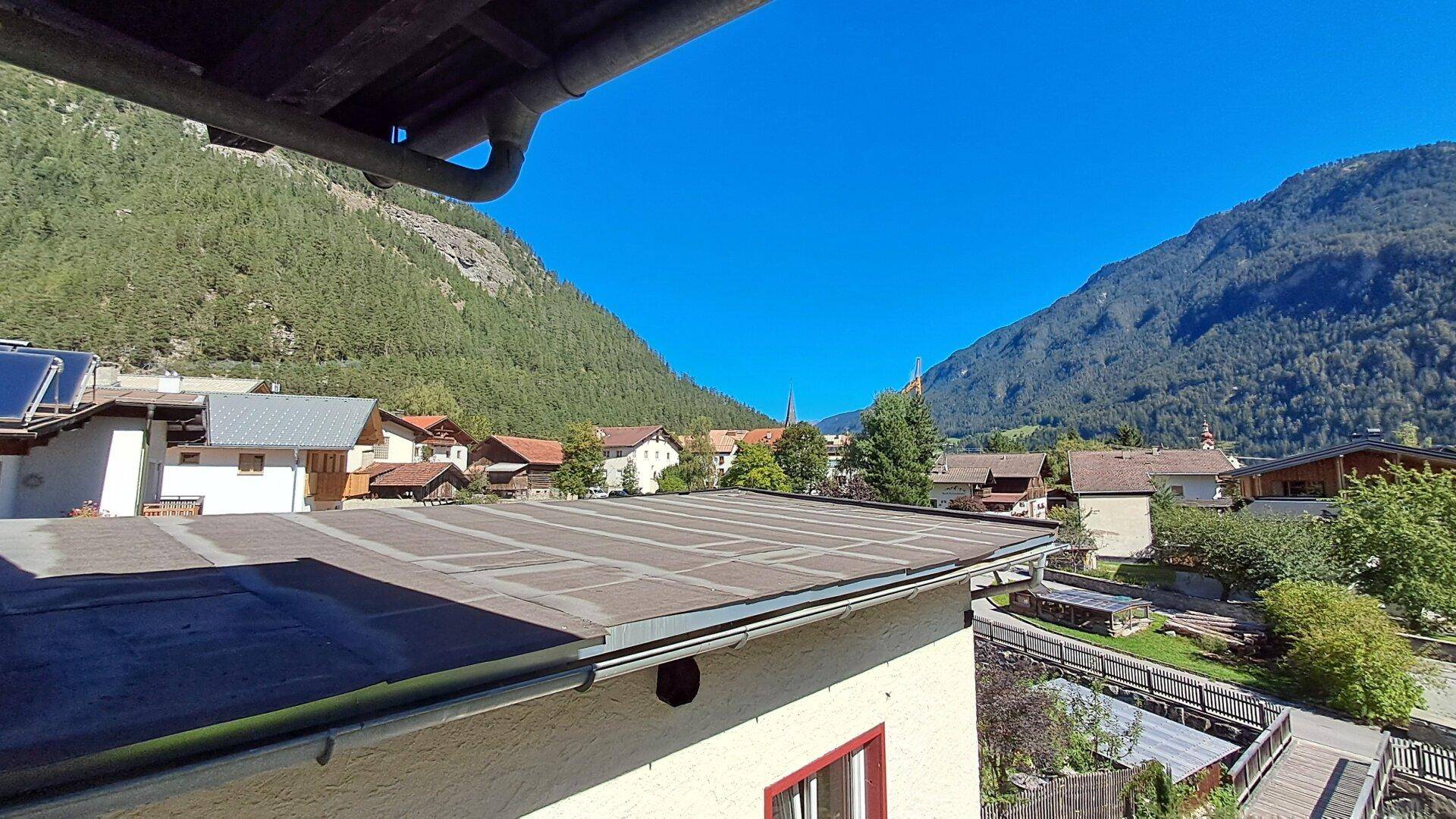 Blick über die Dächer eines Dorfes auf die umliegenden bewaldeten Berge unter strahlend blauem Himmel.
