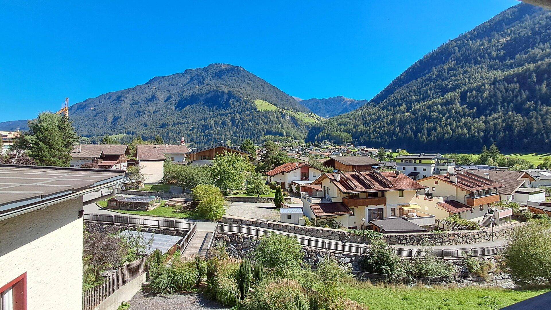 Panoramablick auf ein malerisches Dorf in einem Tal, umgeben von grünen Bergen unter blauem Himmel.