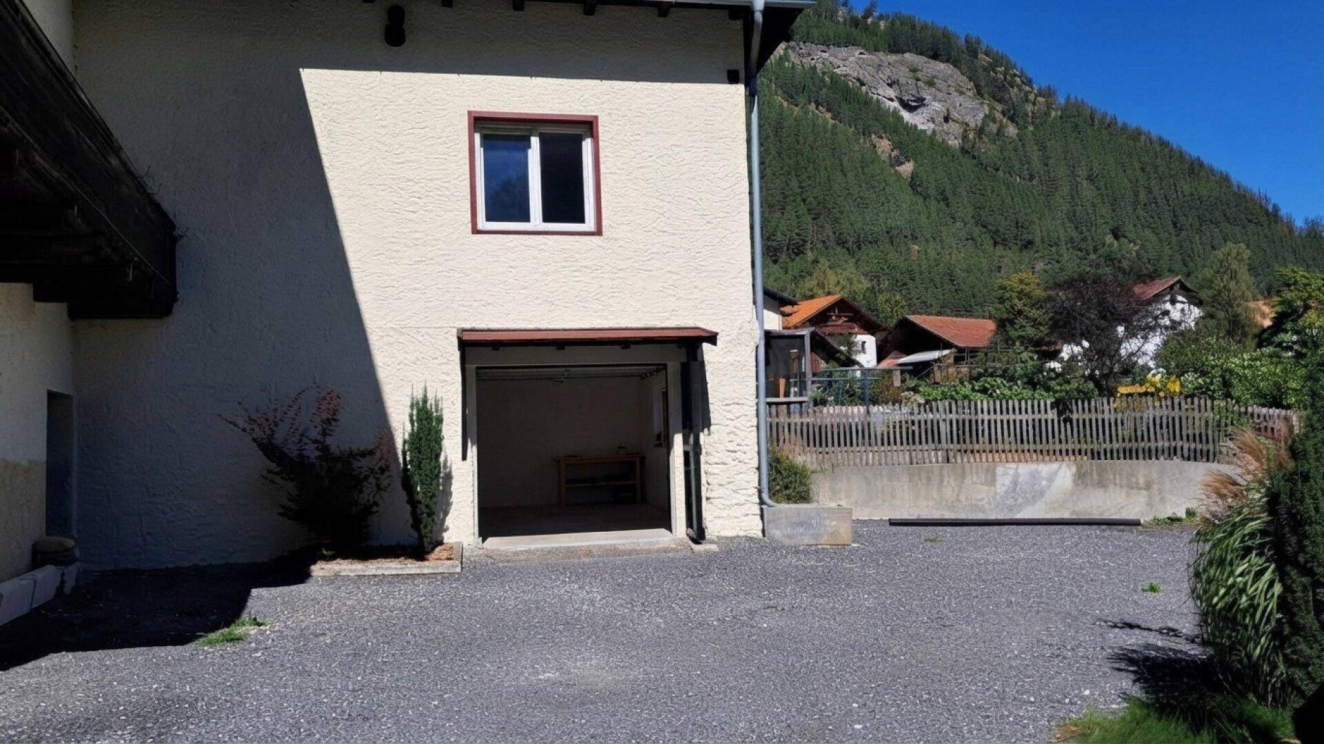 Gebäudeseite mit offener Garage und Schotterfläche, umgeben von grünen Hügeln und Bergen.