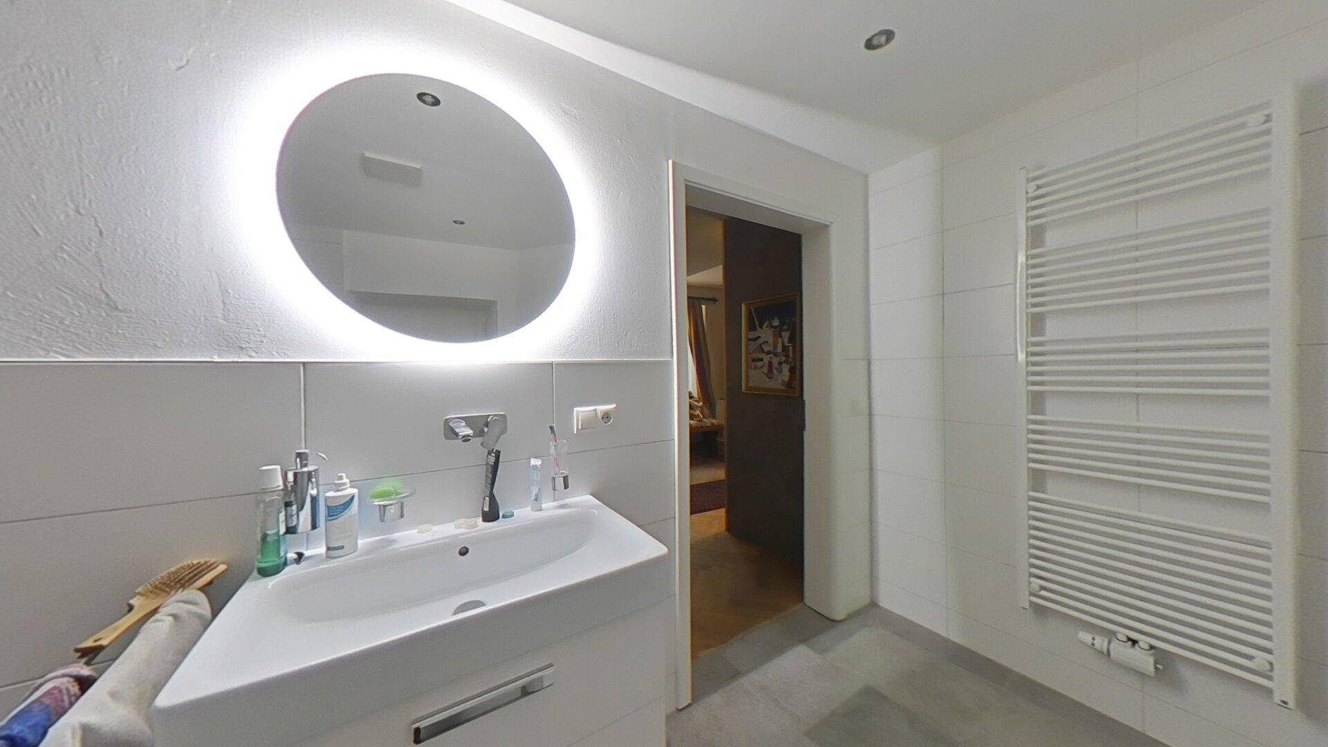 Badezimmer mit einem einzelnen Waschbecken, einem beleuchteten Spiegel und einem Handtuchheizkörper.