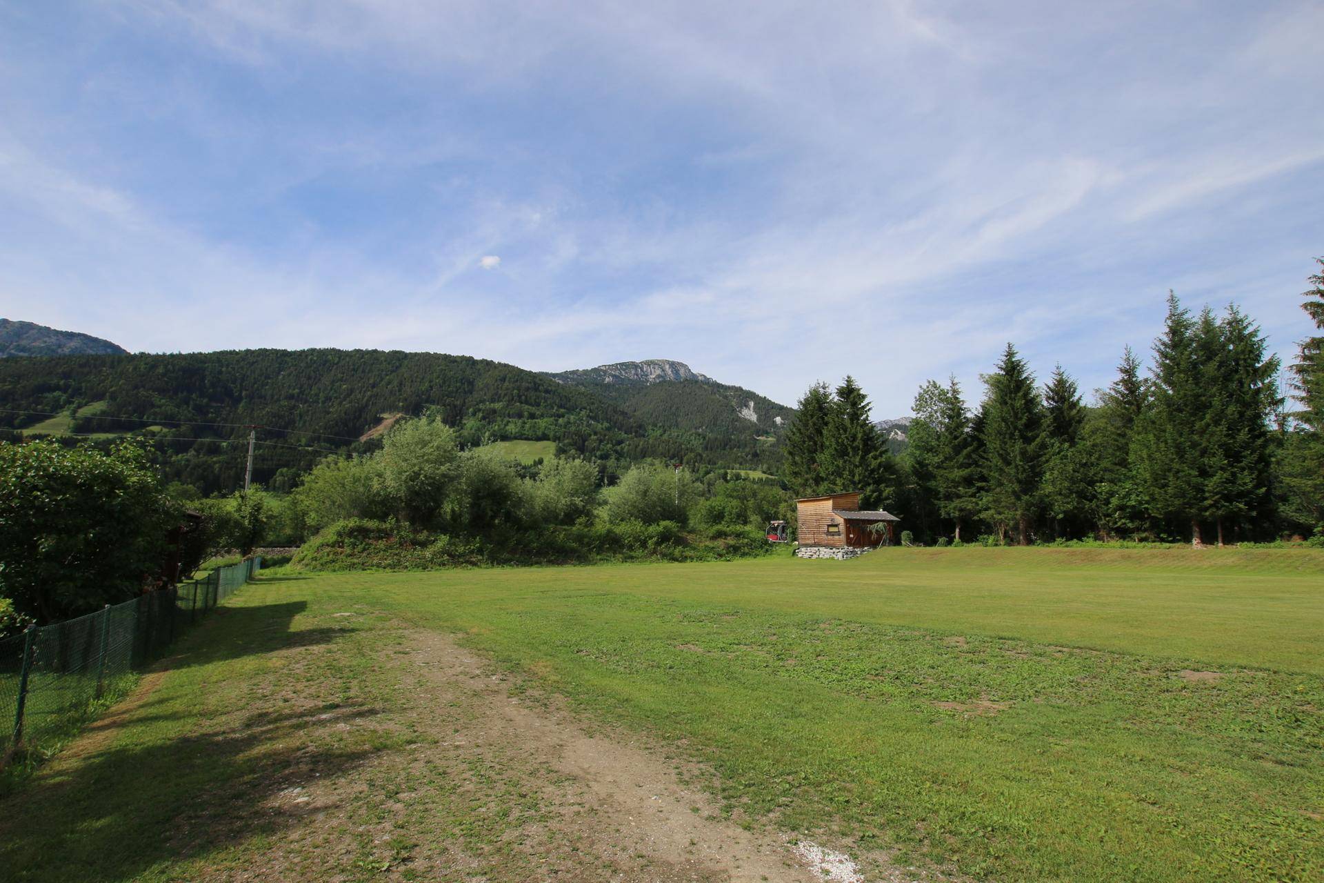 Weite grüne Fläche mit Bäumen und einem kleinen Holzgebäude, umrahmt von bewaldeten Bergen unter blauem Himmel.
