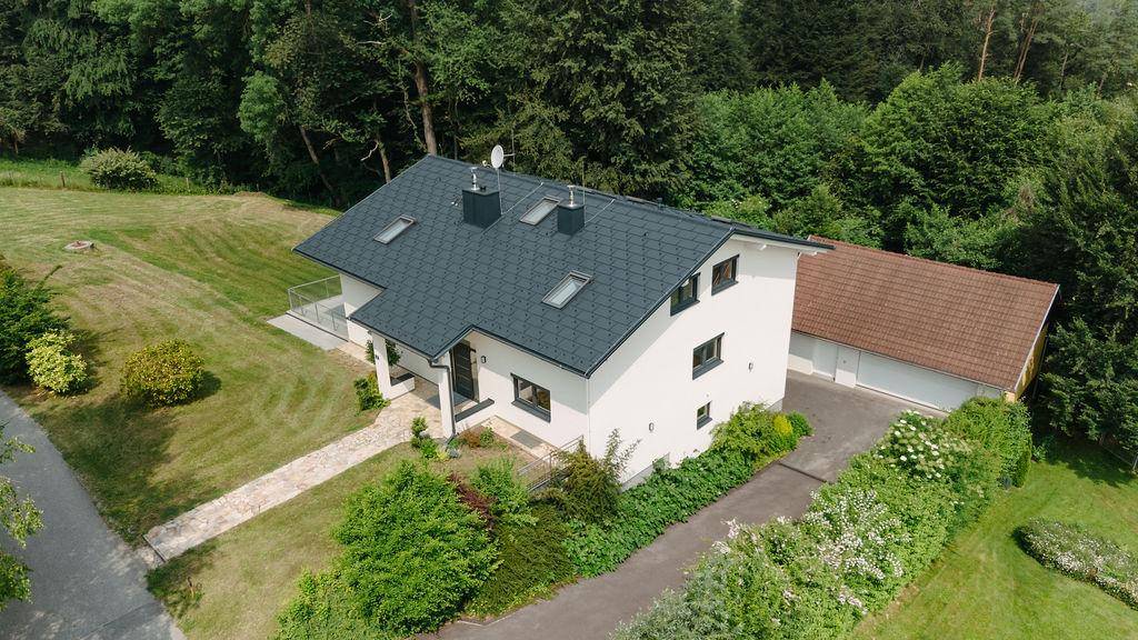 Luftaufnahme eines modernen Hauses mit dunkelgrauem Dach, umgeben von Grünflächen und Bäumen.