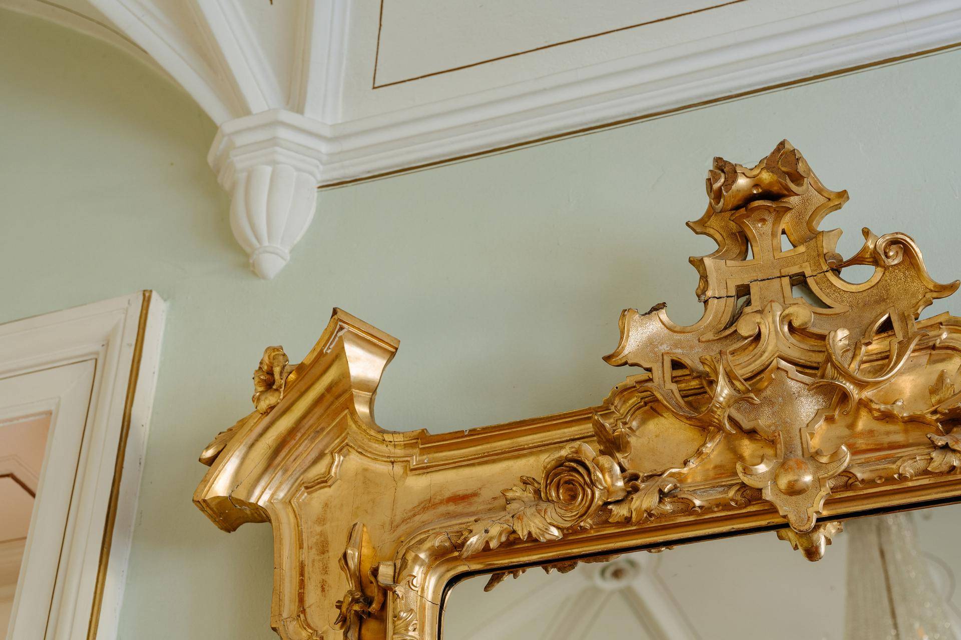 Detail eines opulenten, goldgerahmten Spiegels an einer hellgrünen Wand.