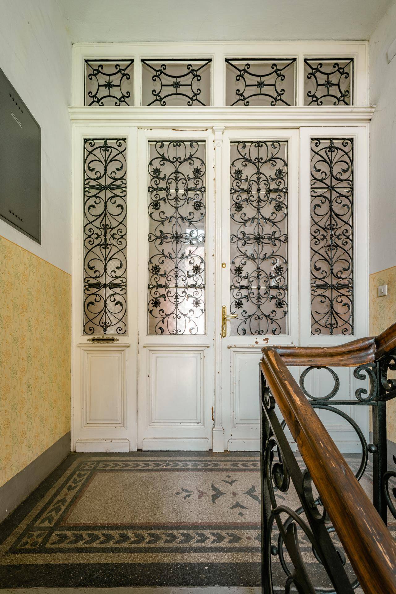 Heller Eingangsbereich mit verzierter Glastür, Terrazzoboden und historischem Treppengeländer.