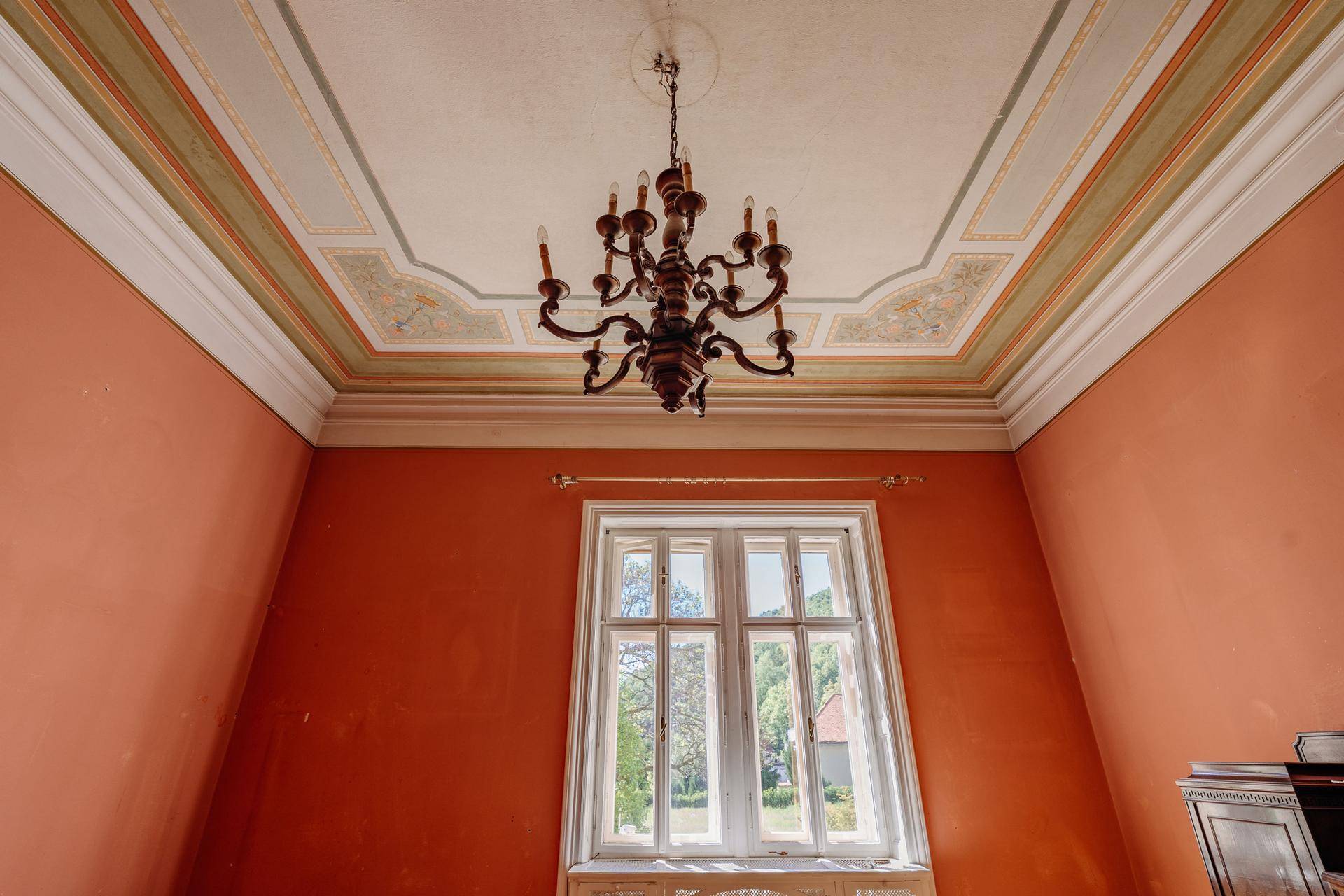 Innenraum mit hohen Decken, Stuckverzierung und einem Kronleuchter, durch ein Fenster fällt Licht.