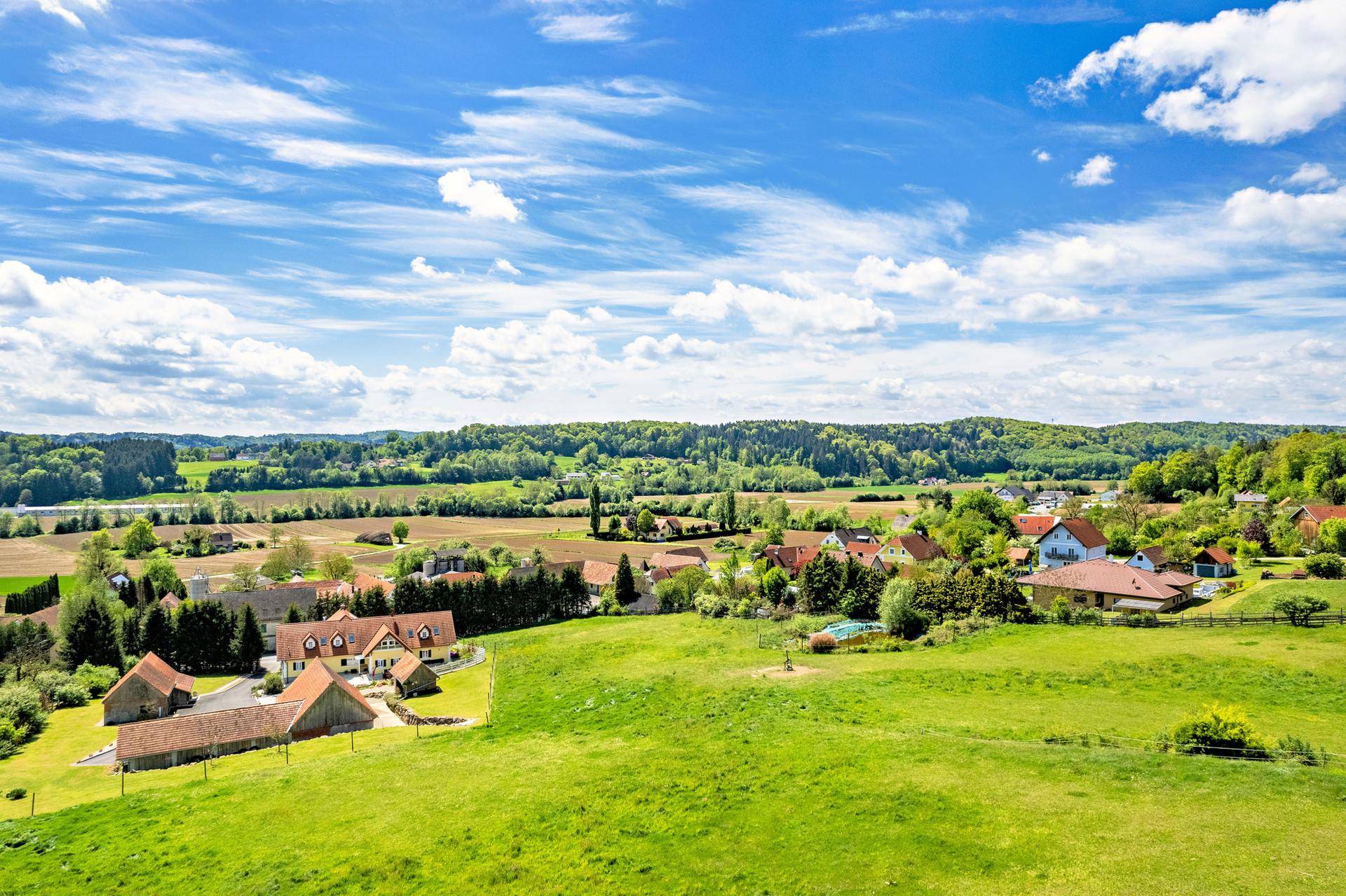 Idyllische ländliche Umgebung mit grünen Wiesen, verstreuten Häusern und bewaldeten Hügeln unter blauem Himmel.