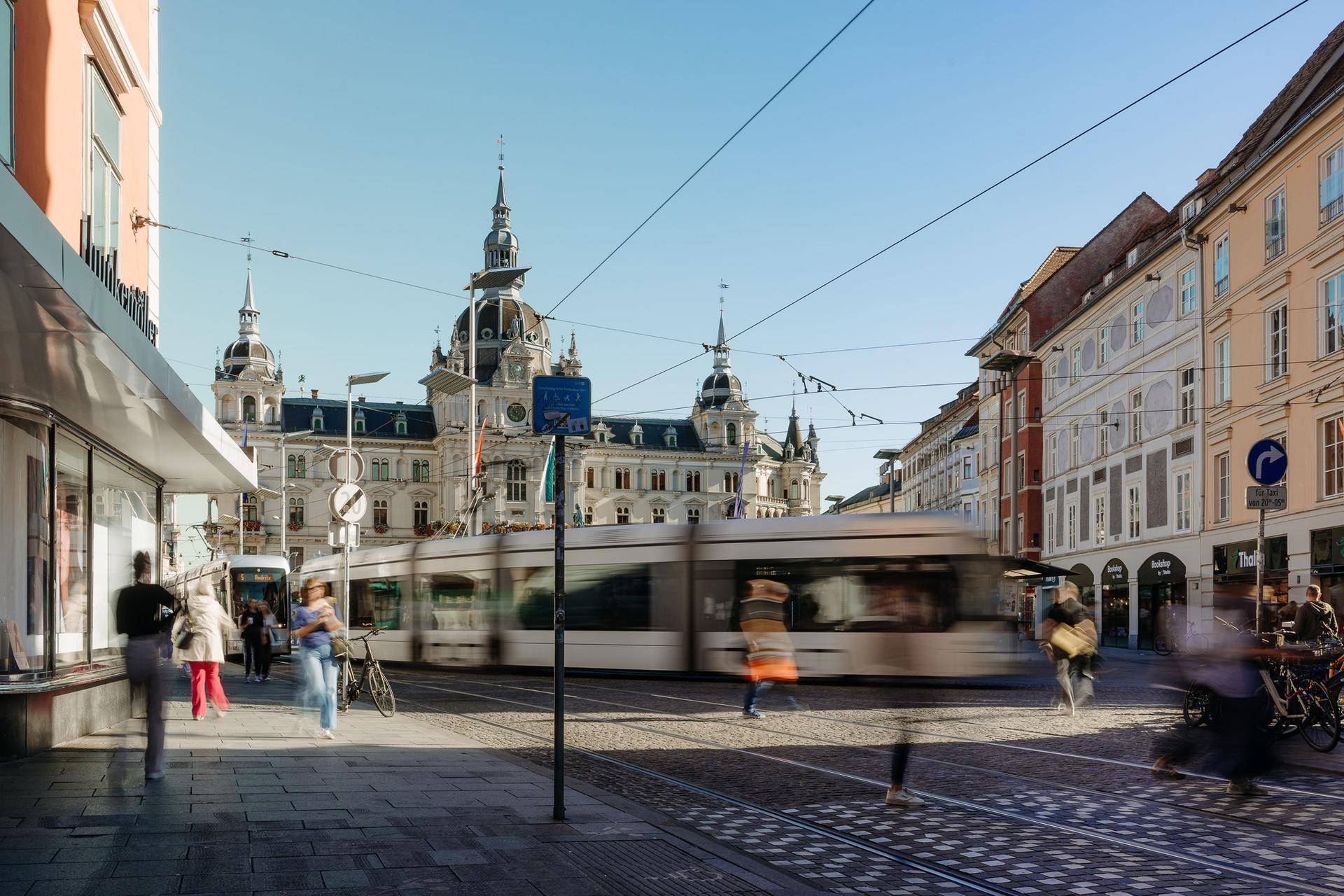 Weitläufige Stadtansicht mit einer Straßenbahn und dem imposanten Grazer Rathaus unter blauem Himmel.
