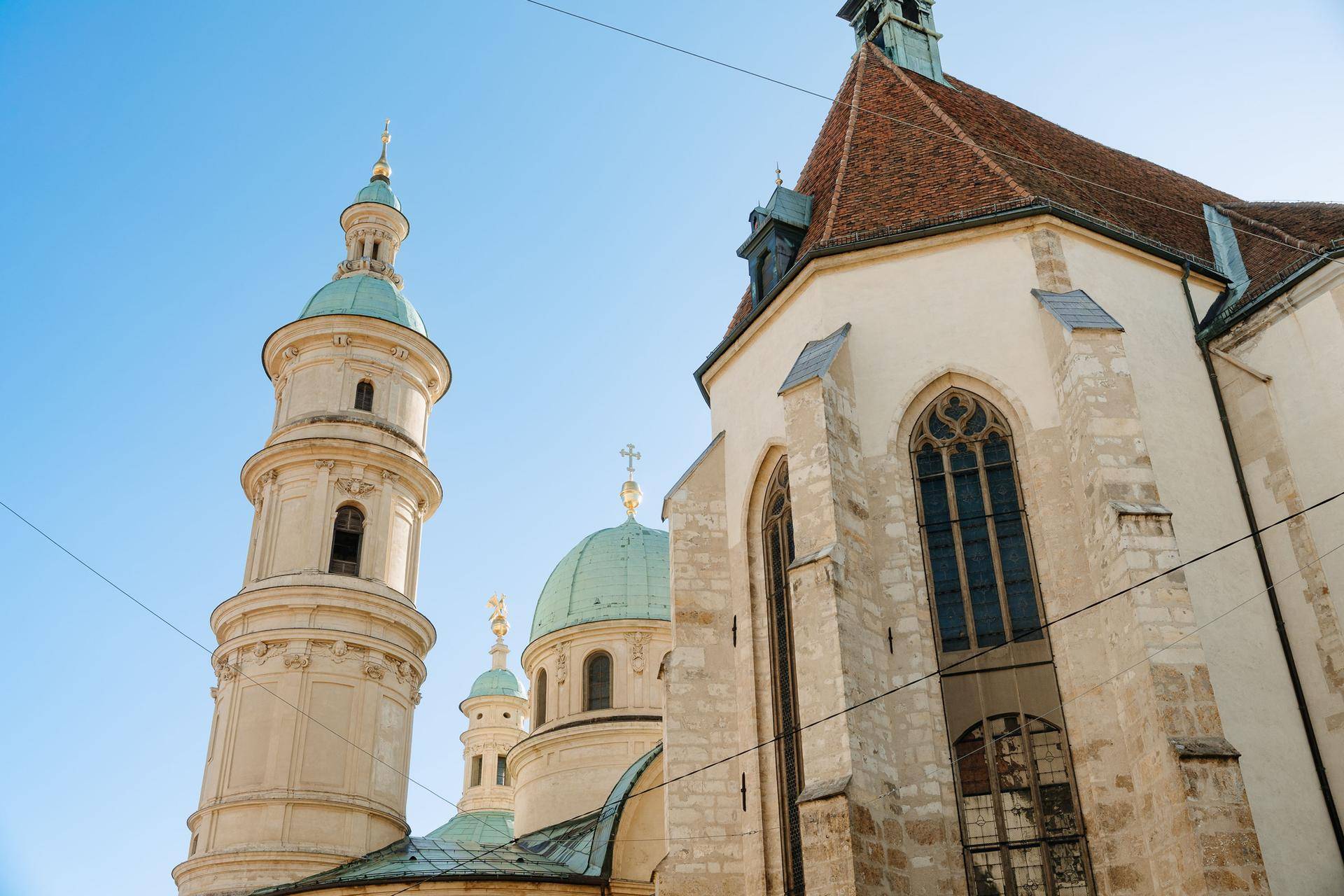 Historische Kirche mit einem hohen Turm, grünen Kuppeln und gotischen Fenstern unter blauem Himmel.