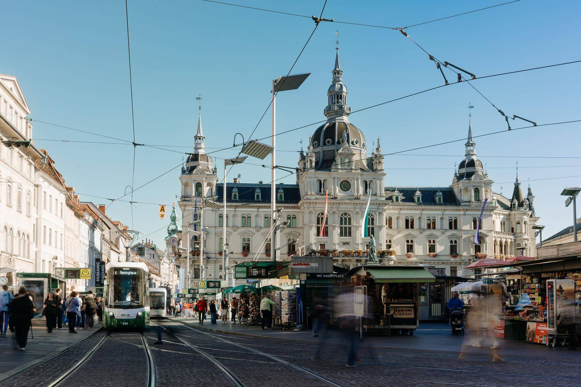 Belebter Stadtplatz mit historischen Gebäuden, Straßenbahnen und Marktständen unter klarem Himmel.