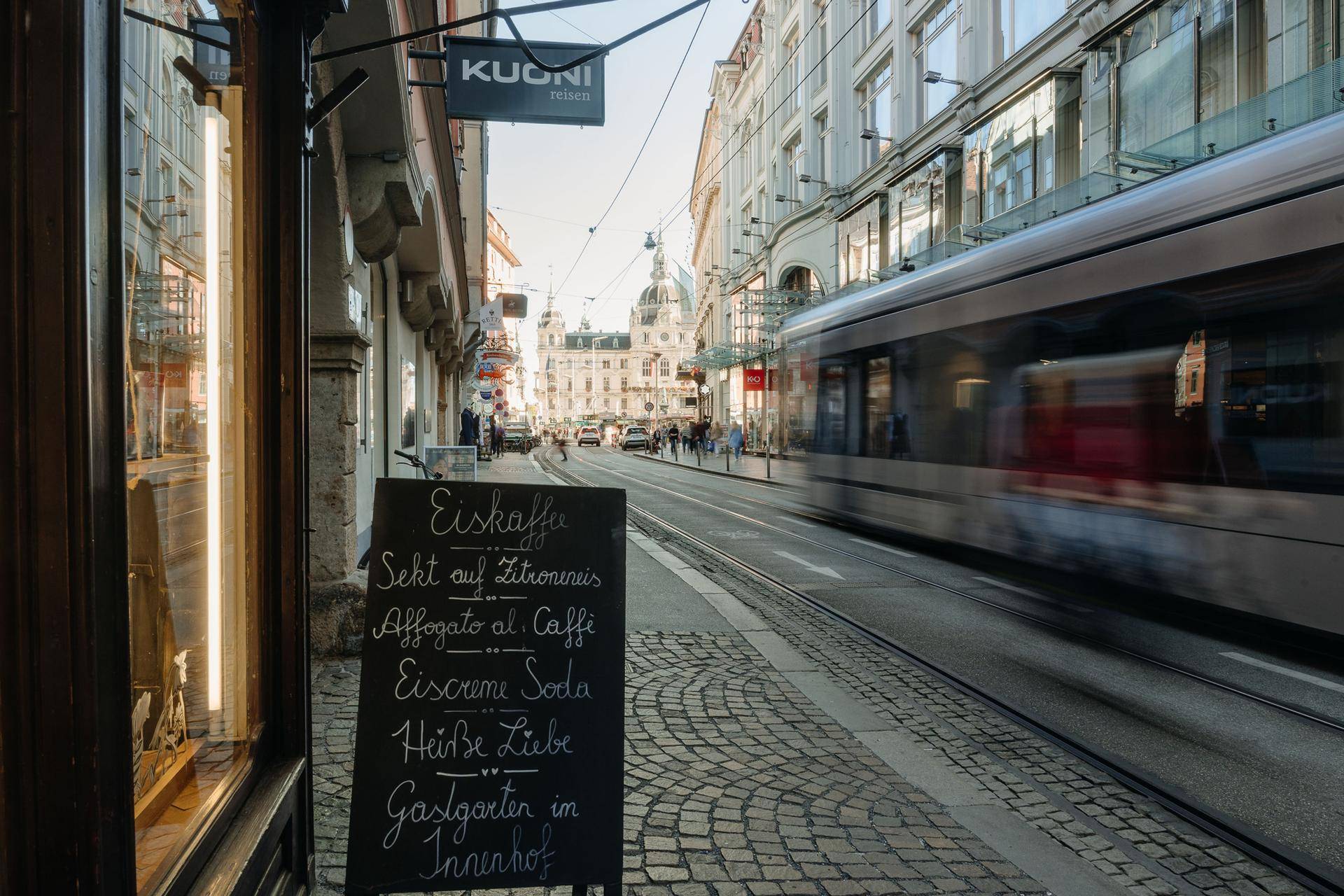 Belebte Stadtstraße mit einer vorbeifahrenden Straßenbahn und einem Kreidetafel-Menü vor einem Geschäft.