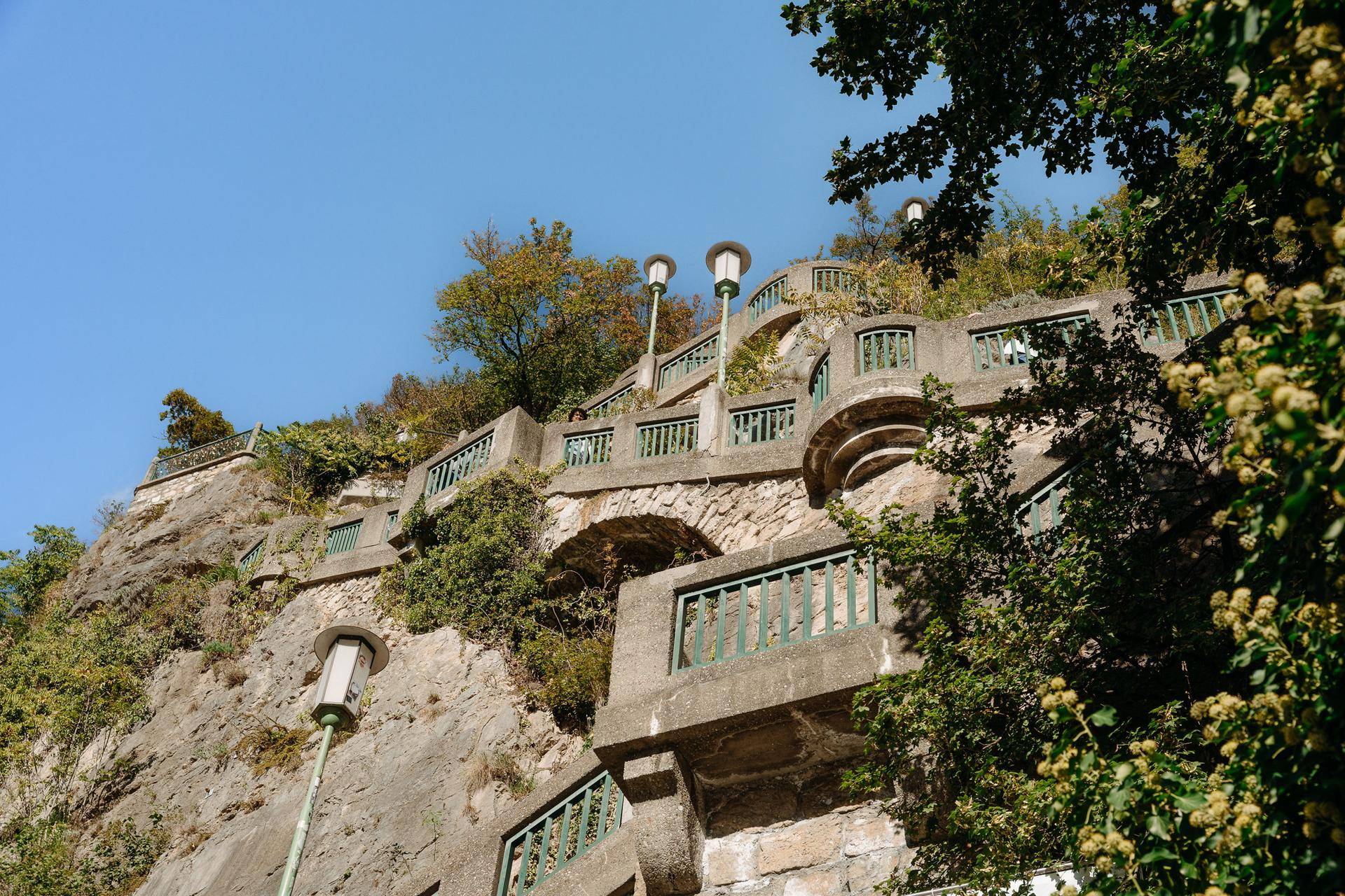 Detailansicht einer alten Steintreppe mit kunstvollen Geländern und Laternen auf einem bewachsenen Felsen.