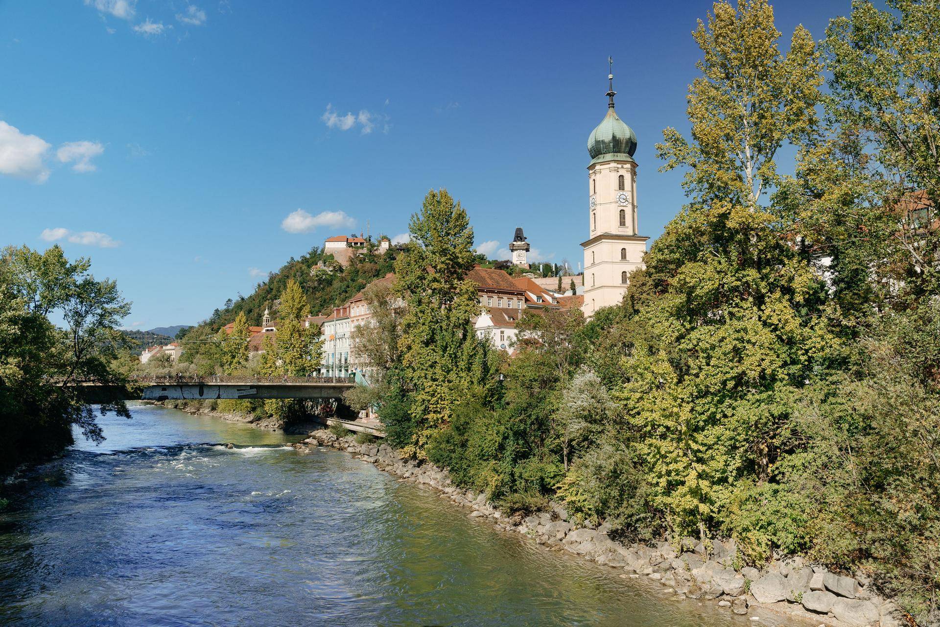 Flusslandschaft mit Brücke, üppigem Grün und historischer Stadtkulisse im Hintergrund.