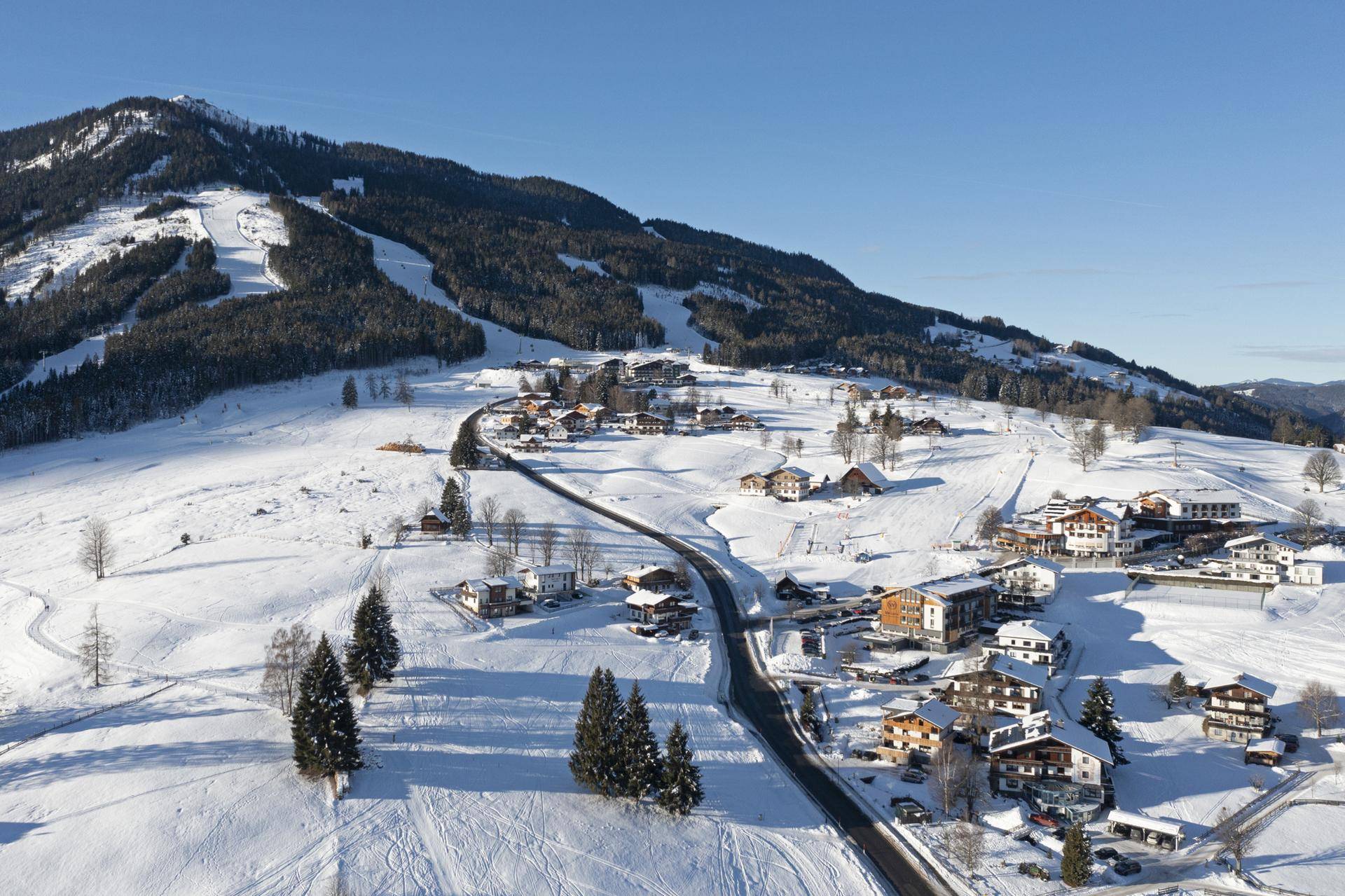 Weitläufiger Winterblick aus der Vogelperspektive auf ein verschneites Dorf mit Skipisten und einer Bergstraße.