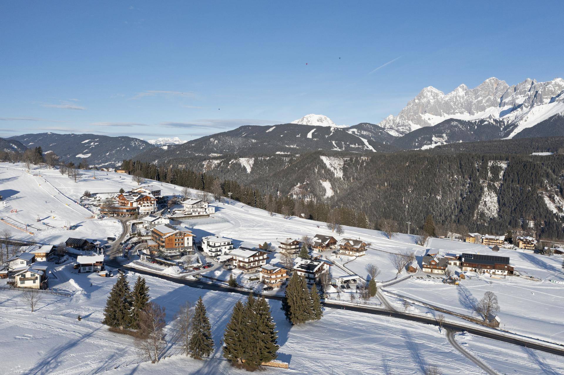 Winterlicher Panoramablick aus der Vogelperspektive auf ein verschneites Dorf, umgeben von schneebedeckten Bergen.