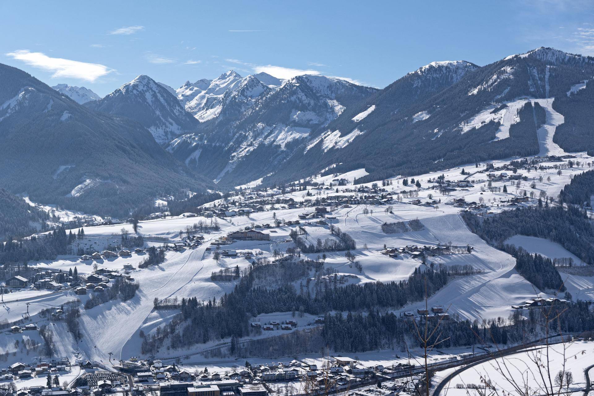 Breiter Panoramablick auf ein verschneites Tal mit einem Dorf, umgeben von majestätischen Bergen unter blauem Himmel.