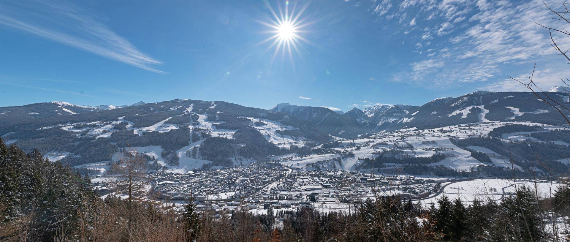 Sonniger Panoramablick auf eine verschneite Berglandschaft mit einem Dorf im Tal unter strahlend blauem Himmel.