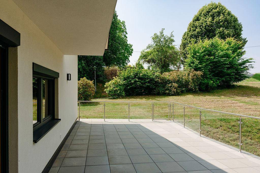 Geräumige, geflieste Terrasse am Haus mit Glasgeländer und Blick ins Grüne.