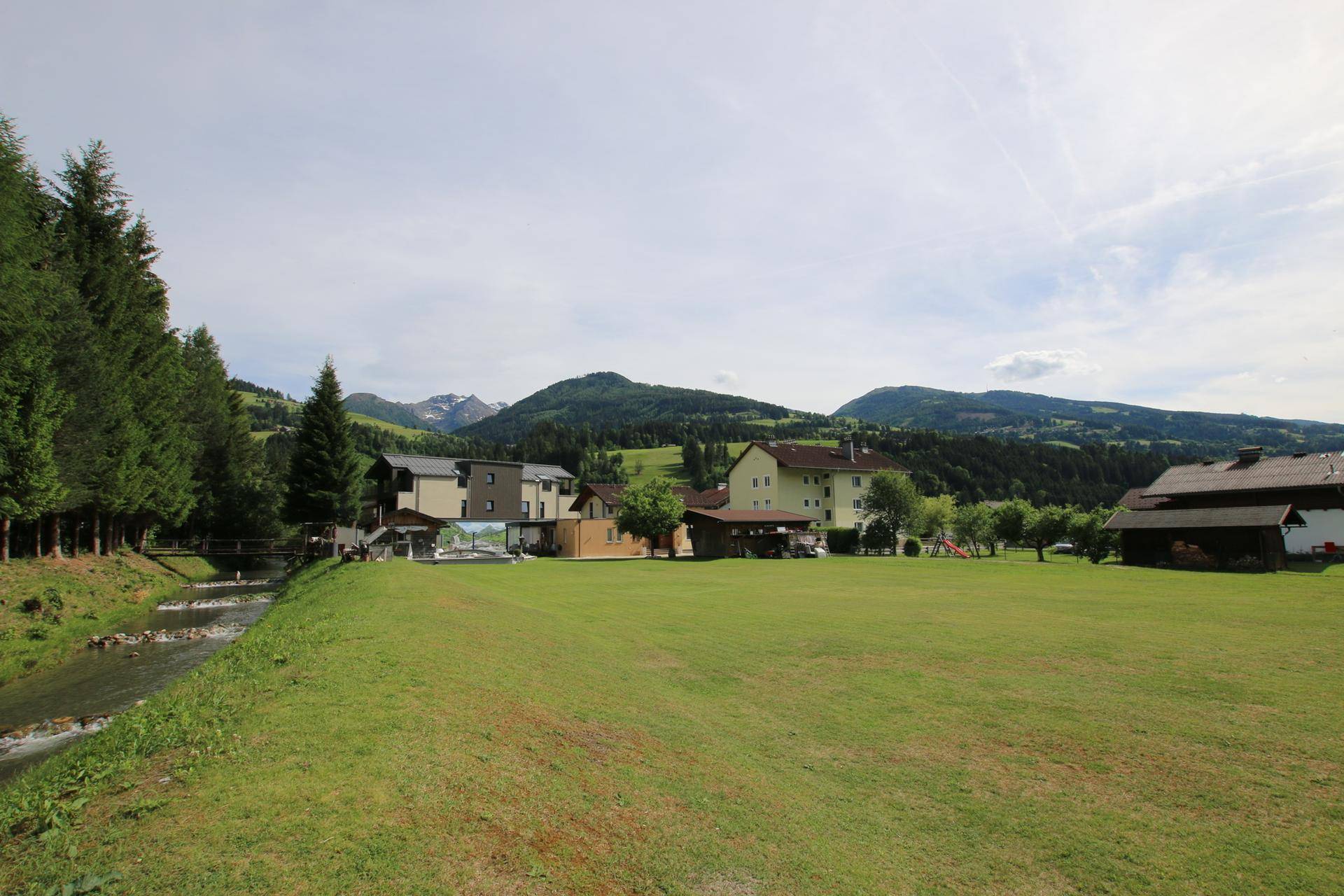 Weitläufige grüne Landschaft mit Gebäuden, einem Bach und majestätischen Bergen im Hintergrund unter blauem Himmel.