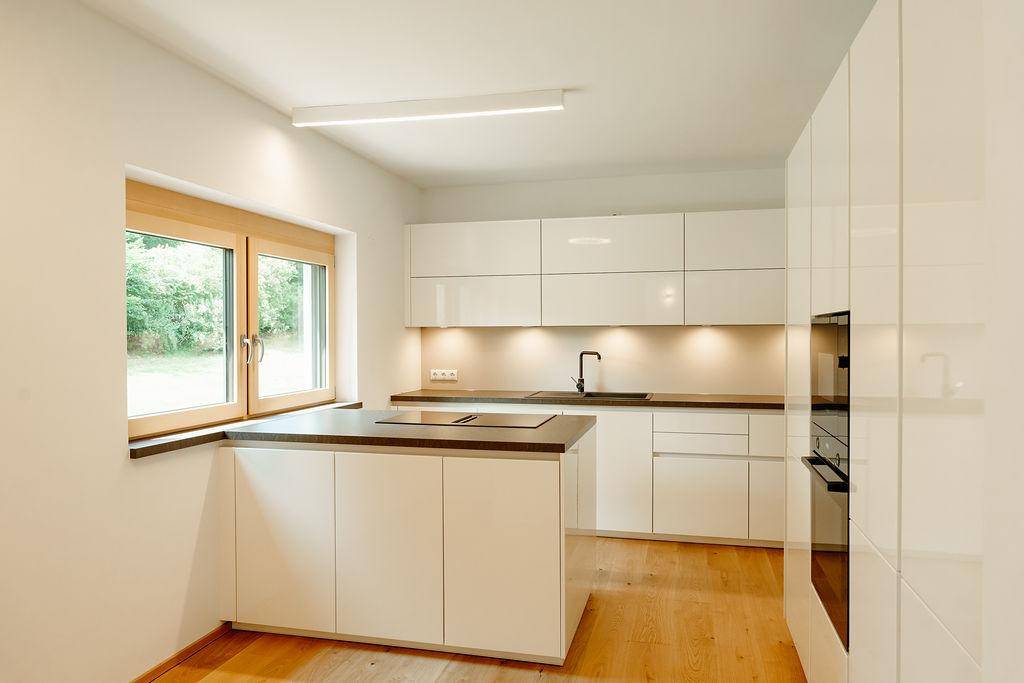 Moderne Küche mit weißen Hochglanzfronten, Holzboden und Blick ins Grüne.