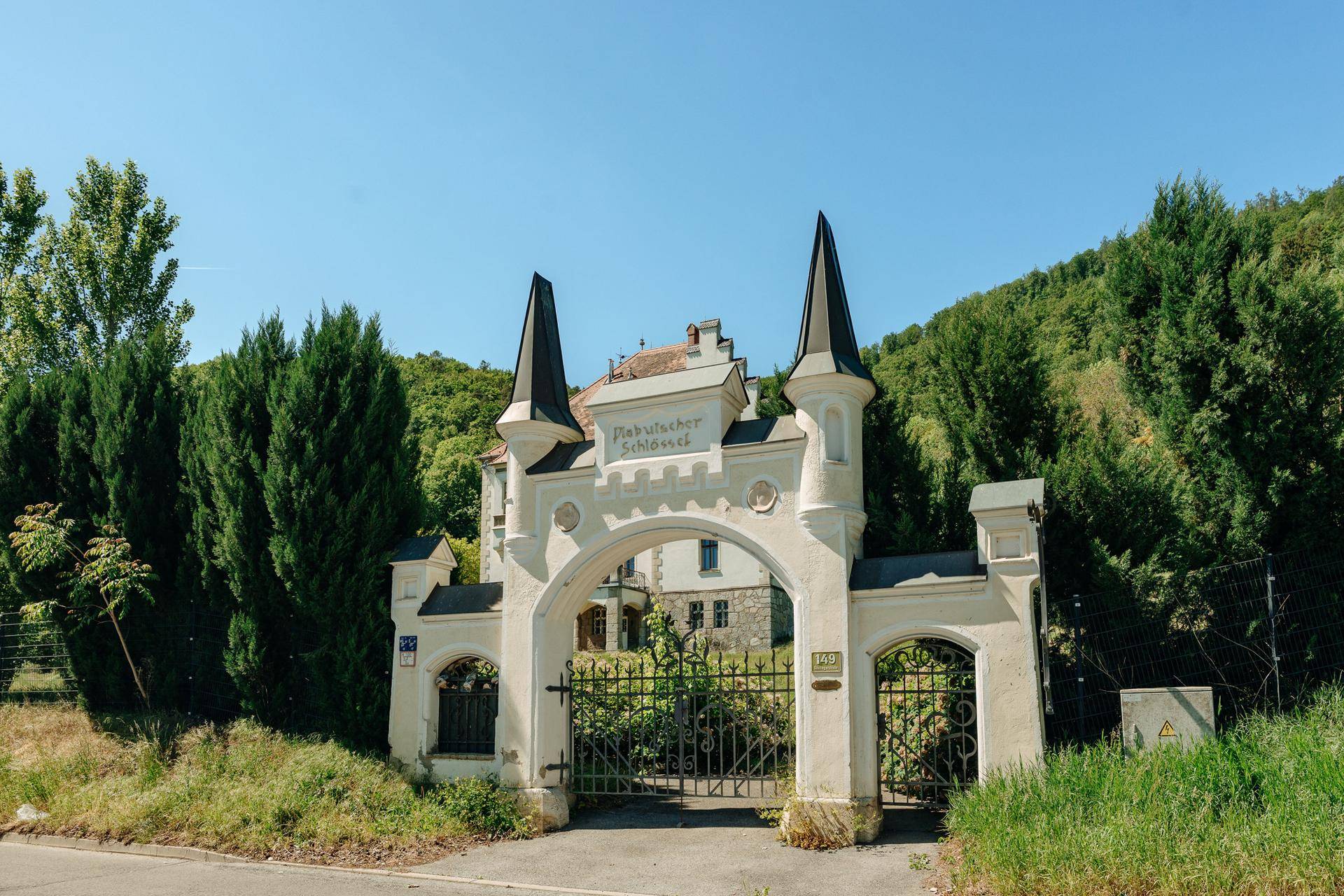 Historisches Eingangstor mit Türmchen und schmiedeeisernen Gittern, umgeben von Bäumen.