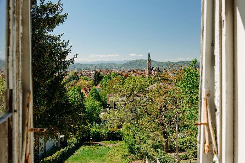 Panoramablick aus einem Fenster auf die Stadt, grüne Bäume und ferne Berge.