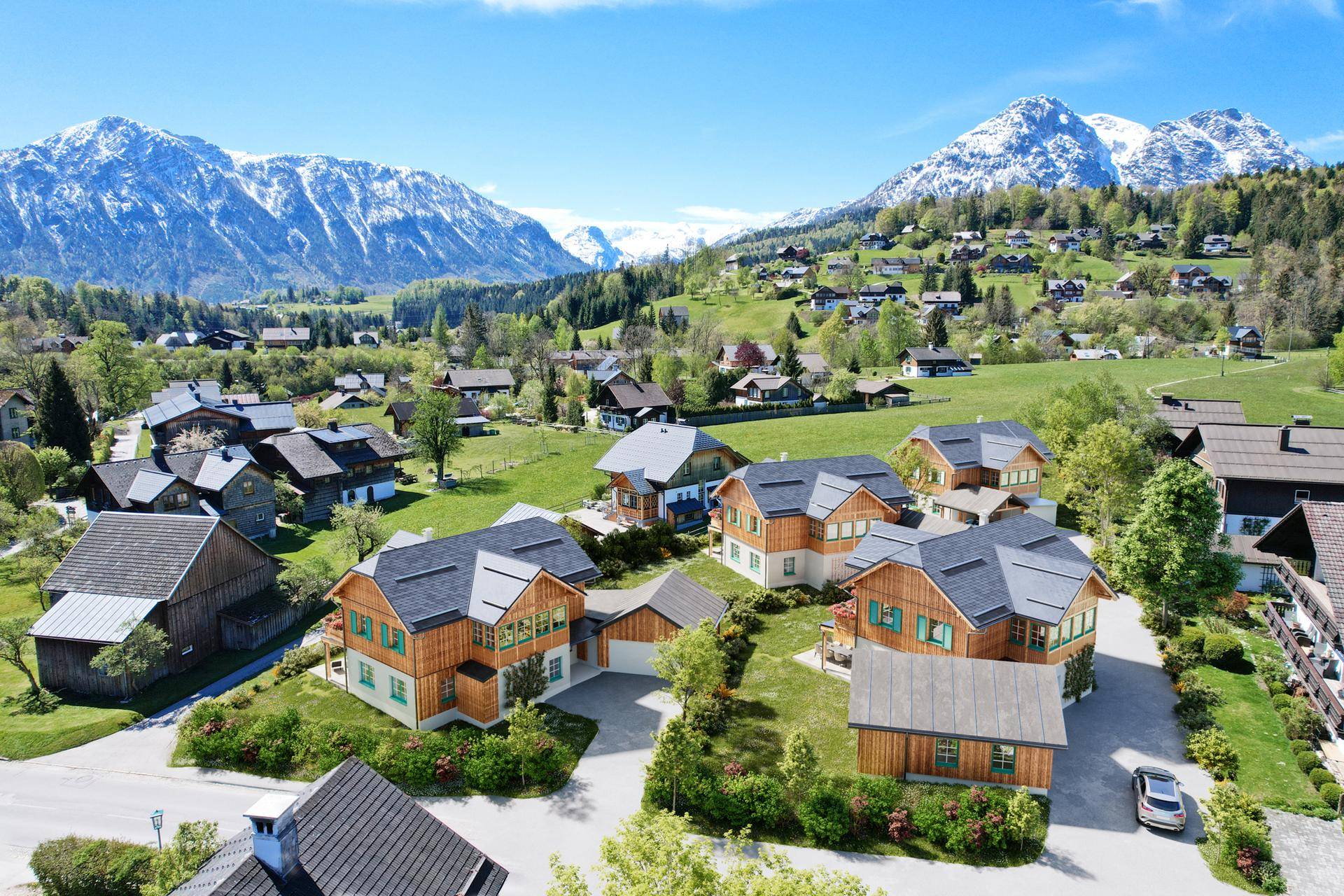 Dorfansicht mit mehreren Häusern und schneebedeckten Bergen im Hintergrund unter blauem Himmel.