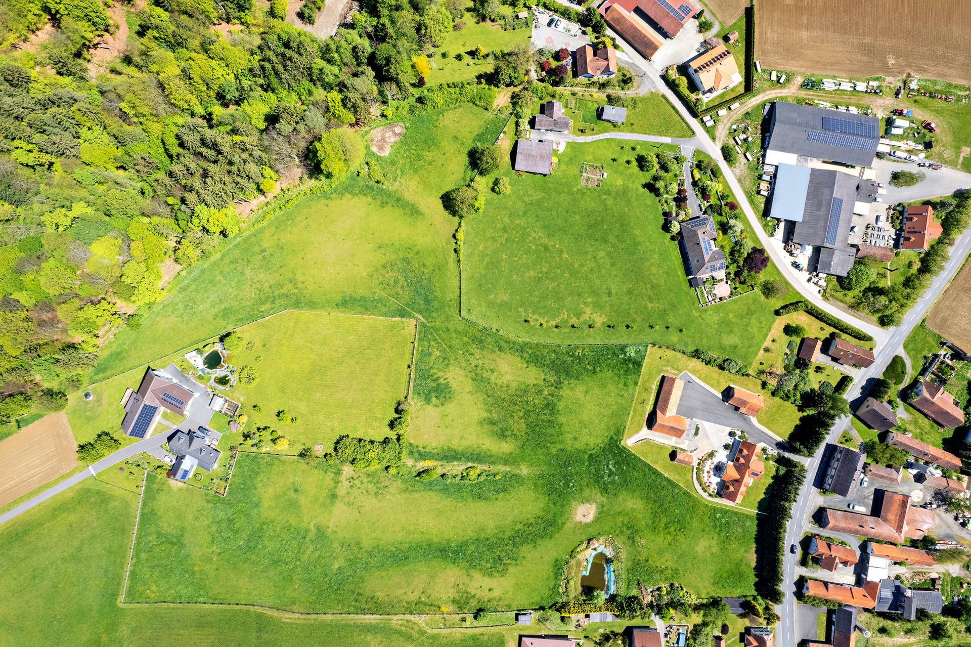 Luftaufnahme eines ländlichen Gebiets mit Feldern, Waldstücken, Wohnhäusern und einer kleinen Straße.