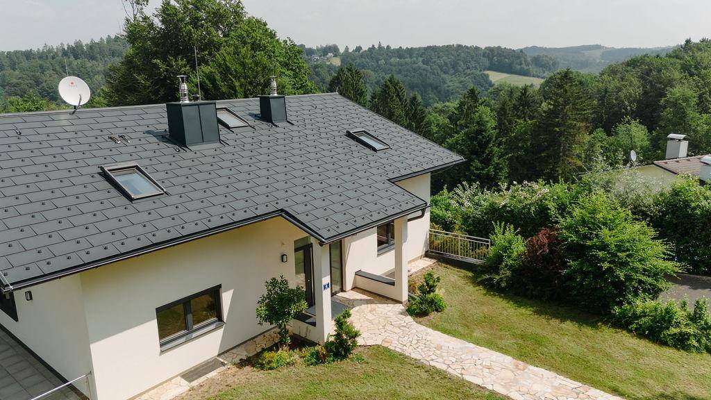 Ansicht des Hauses mit dunkelgrauem Dach und weißer Fassade, umgeben von grüner Hügellandschaft.