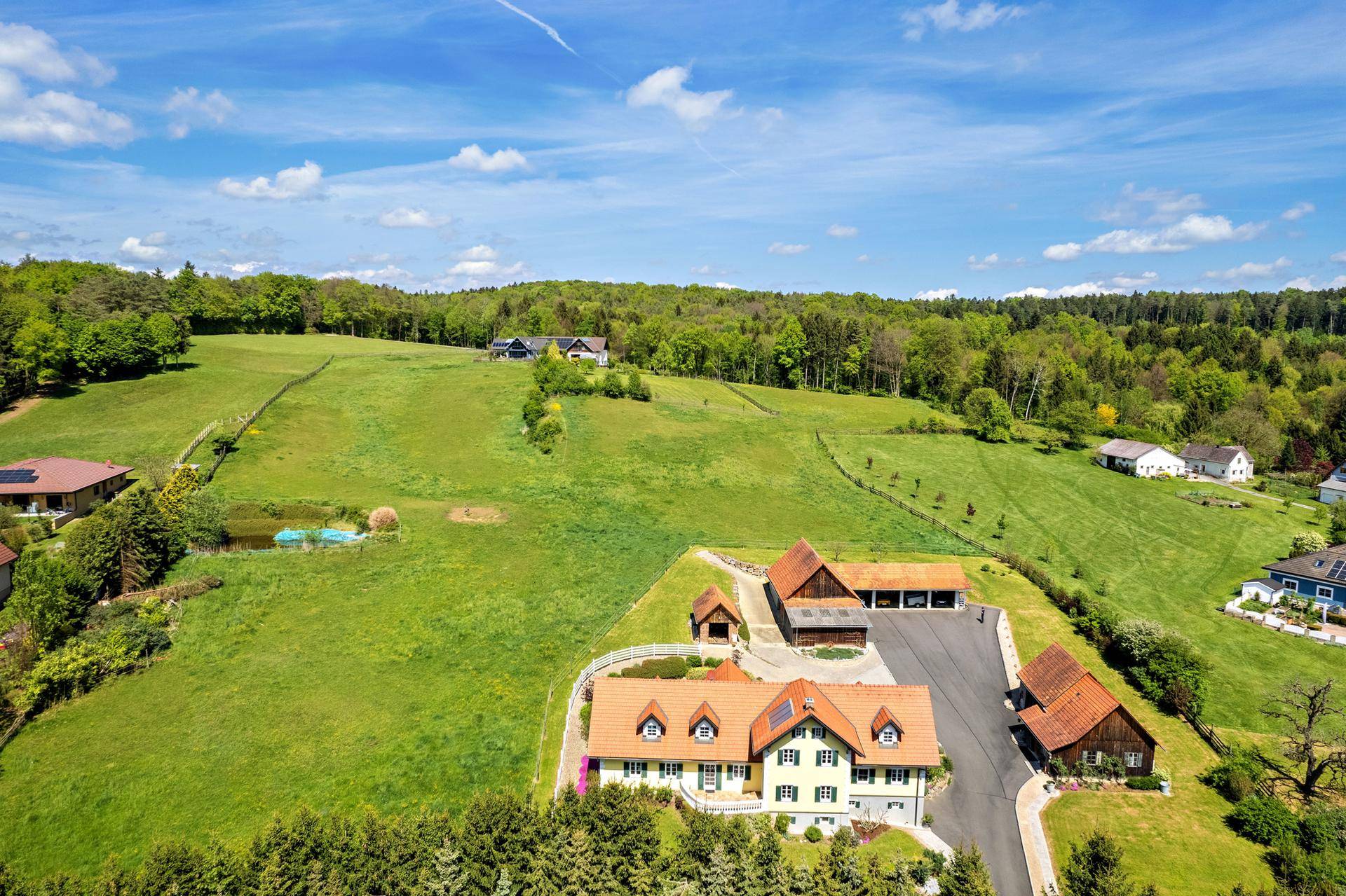Panoramablick auf ein ländliches Anwesen mit mehreren Gebäuden, grünen Wiesen und bewaldeten Hügeln.