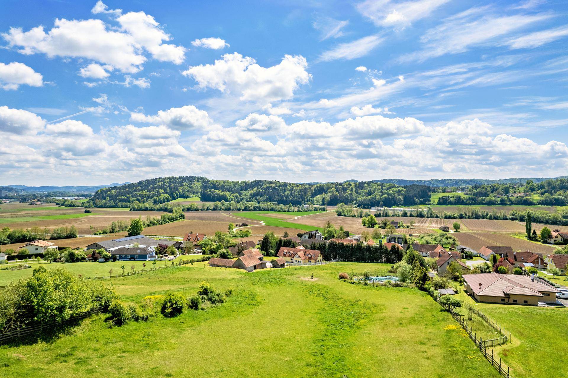 Weitläufige Landschaft mit grünen Feldern, vereinzelten Häusern und bewaldeten Hügeln unter blauem Himmel.