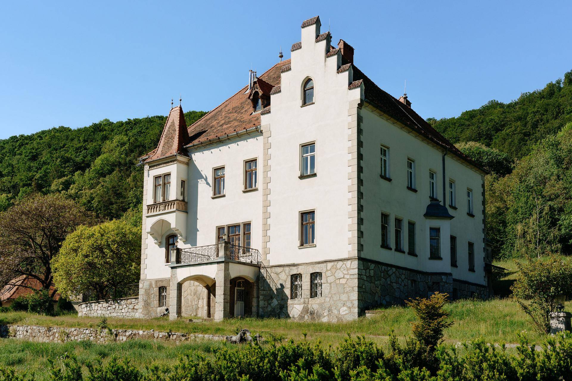 Historisches Gebäude mit weißer Fassade und rotem Ziegeldach, umgeben von grüner Natur.
