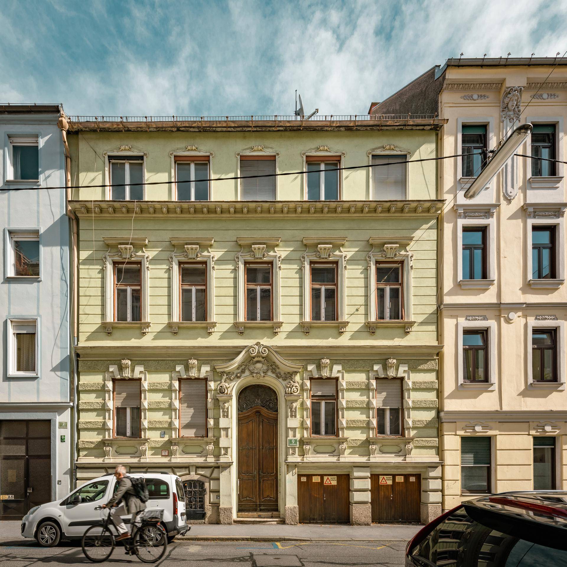 Historisches Gebäude mit grüner Fassade, verzierten Details und Rollläden an einer Stadtstraße.
