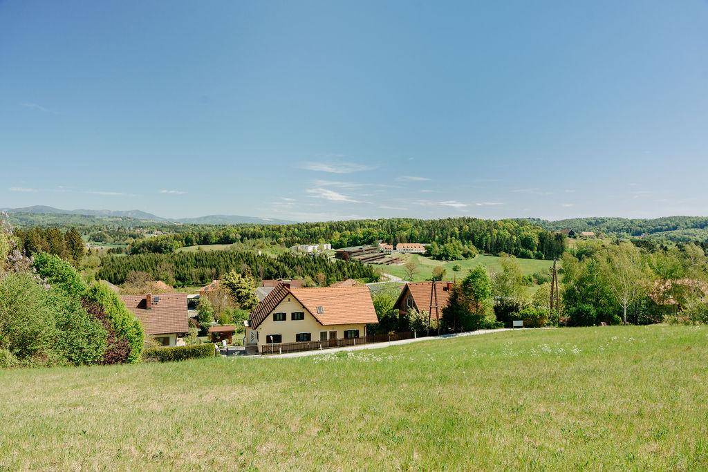 Panoramablick über eine grüne Wiese auf eine kleine Siedlung und die umliegende bewaldete Hügellandschaft.