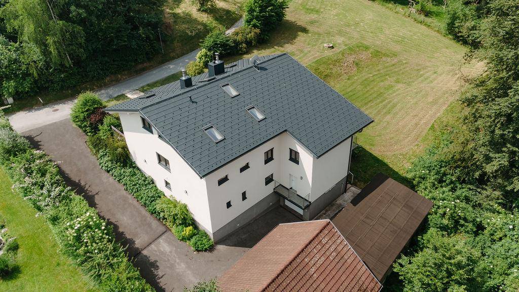 Vogelperspektive des Hauses mit dunkelgrauem Dach und weißer Fassade, eingebettet in grüne Landschaft.