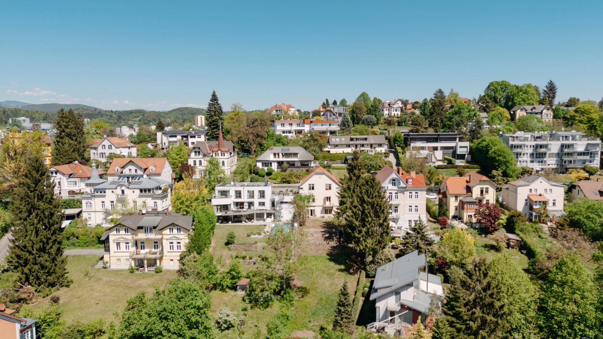 Weitläufige Luftaufnahme einer grünen Wohnsiedlung mit verschiedenen Häusern und viel Baumbestand unter blauem Himmel.