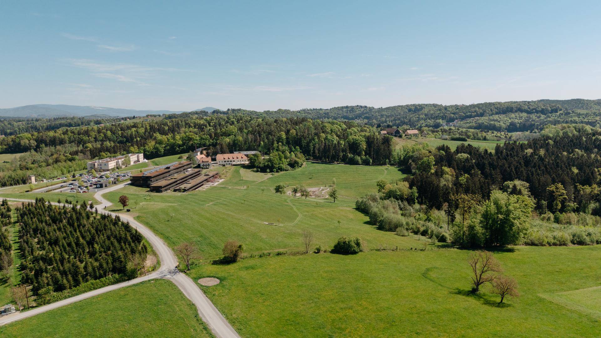 Weitläufige Landschaftsaufnahme mit grünen Feldern, Wäldern und einigen Gebäuden in der Ferne unter blauem Himmel.