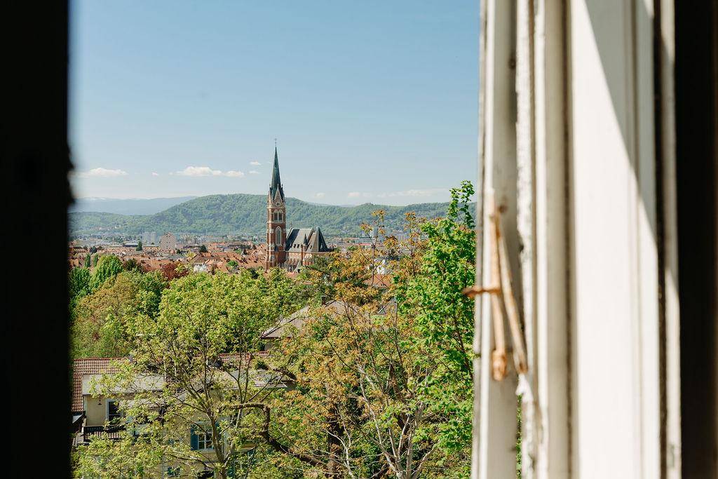 Blick aus einem alten Fensterrahmen auf die Stadt, grüne Landschaft und den Kirchturm.