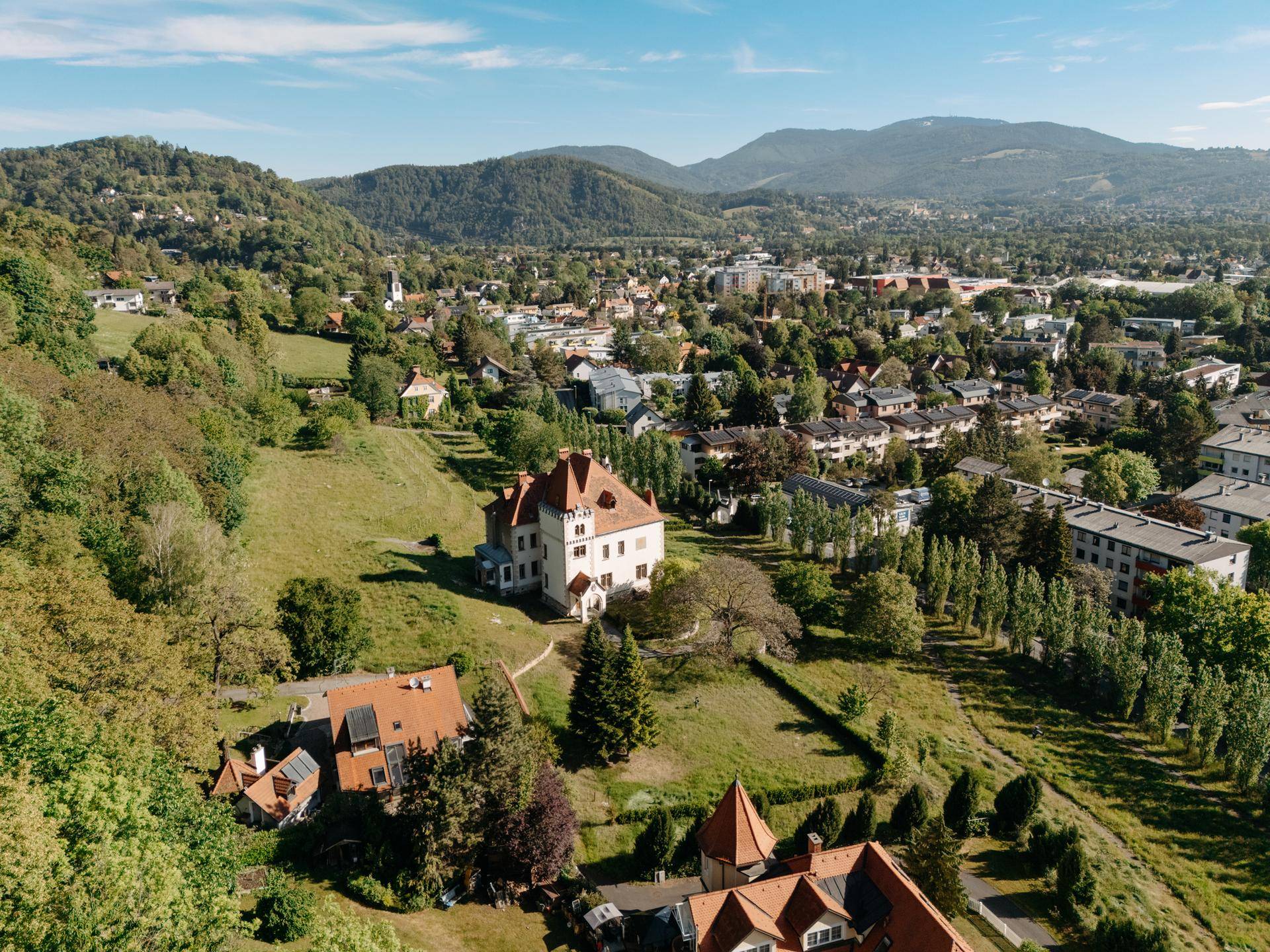 Luftaufnahme des Anwesens in einer grünen Hügellandschaft mit umliegender Bebauung.