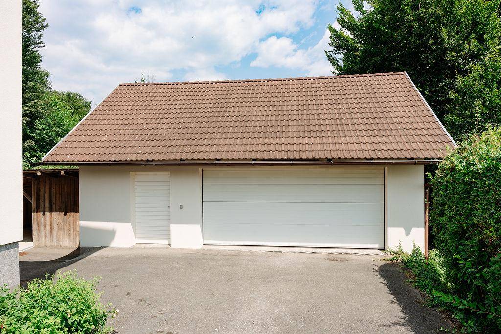 Freistehende Garage mit braunem Ziegeldach, weißem Garagentor und umgebendem Grün.