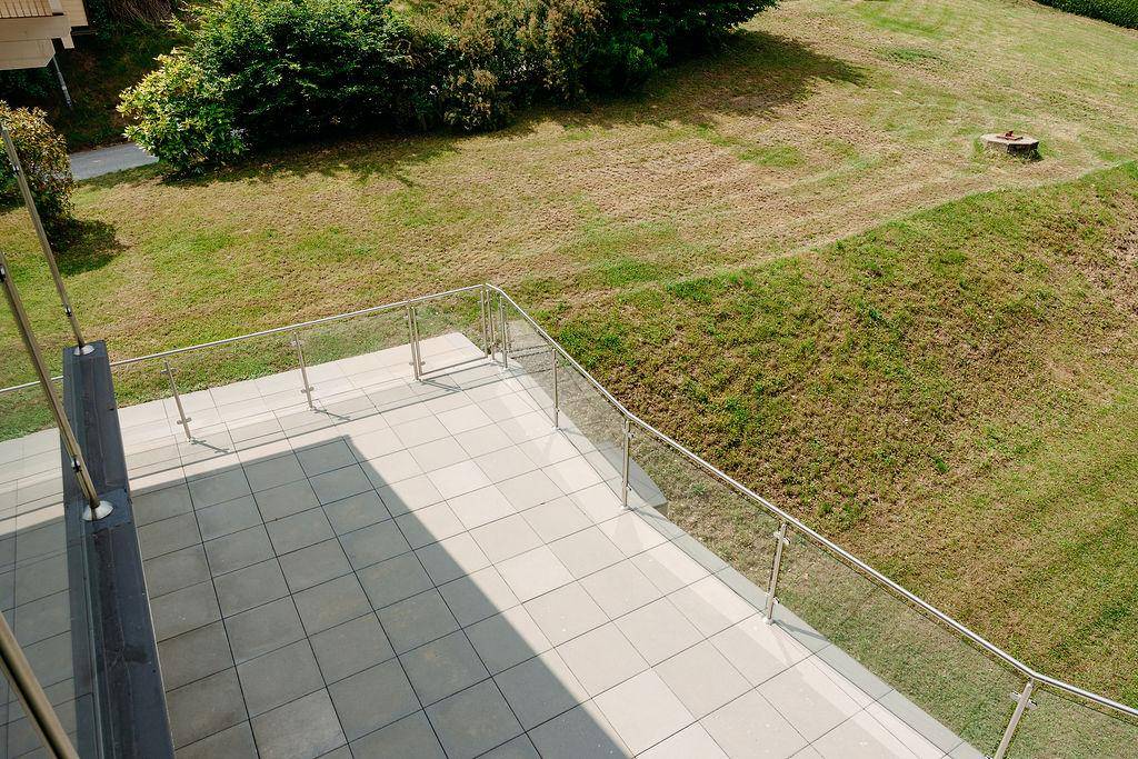 Geräumige Terrasse mit Fliesenboden und Glasgeländer, umgeben von einer Rasenfläche.