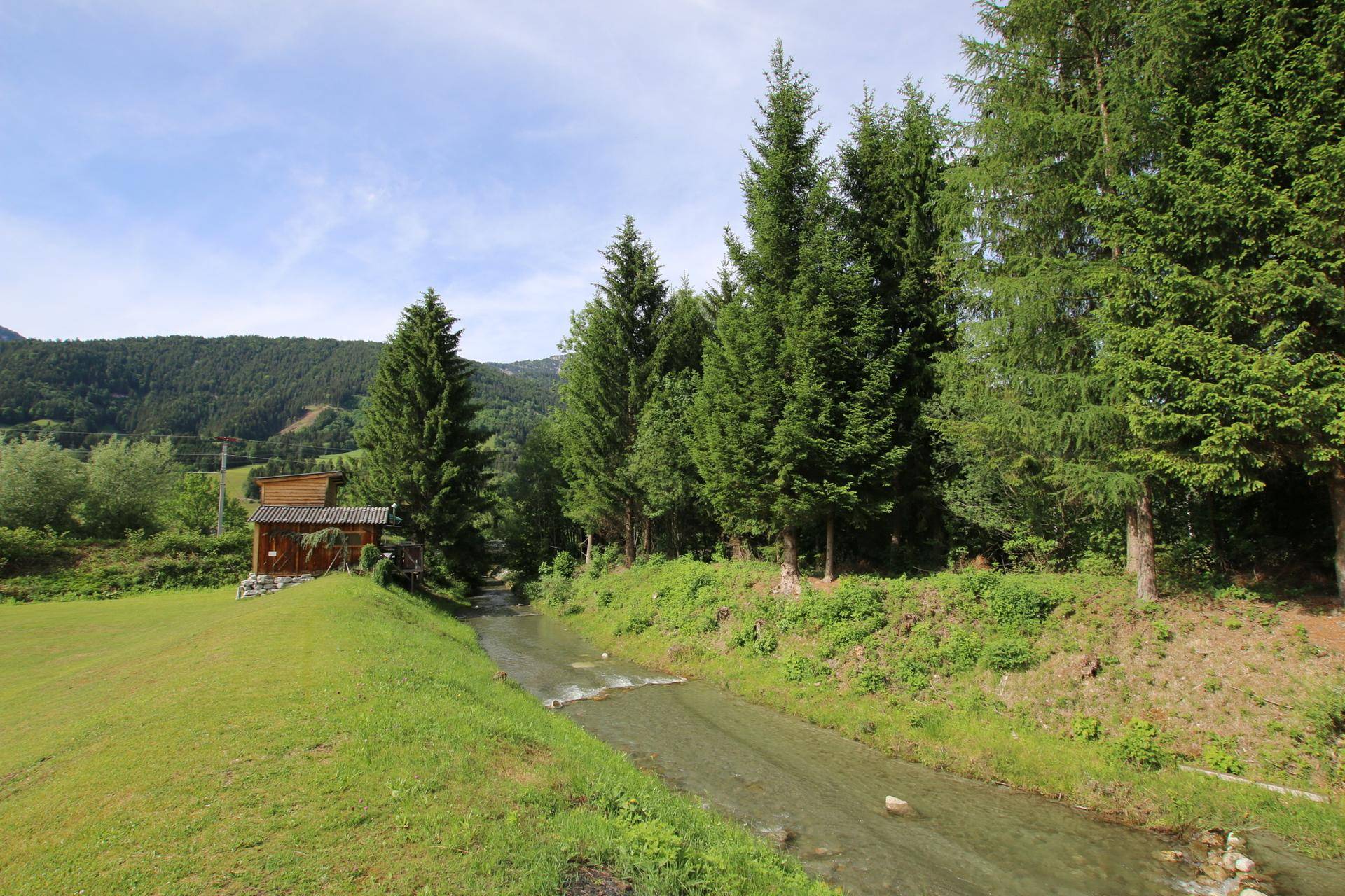 Idyllische Landschaft mit einem Bach, einer weiten Wiese, hohen Bäumen und einem kleinen Holzhaus vor Bergkulisse.