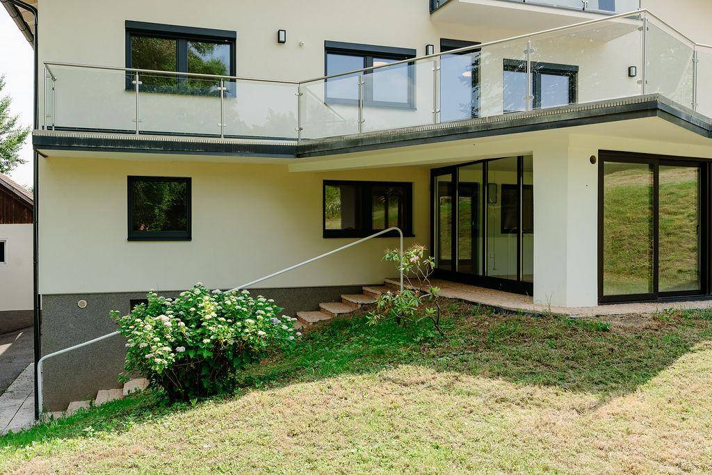 Moderne Hausfassade mit großen Fenstern und Balkon mit Glasgeländer, umgeben von Grünflächen.