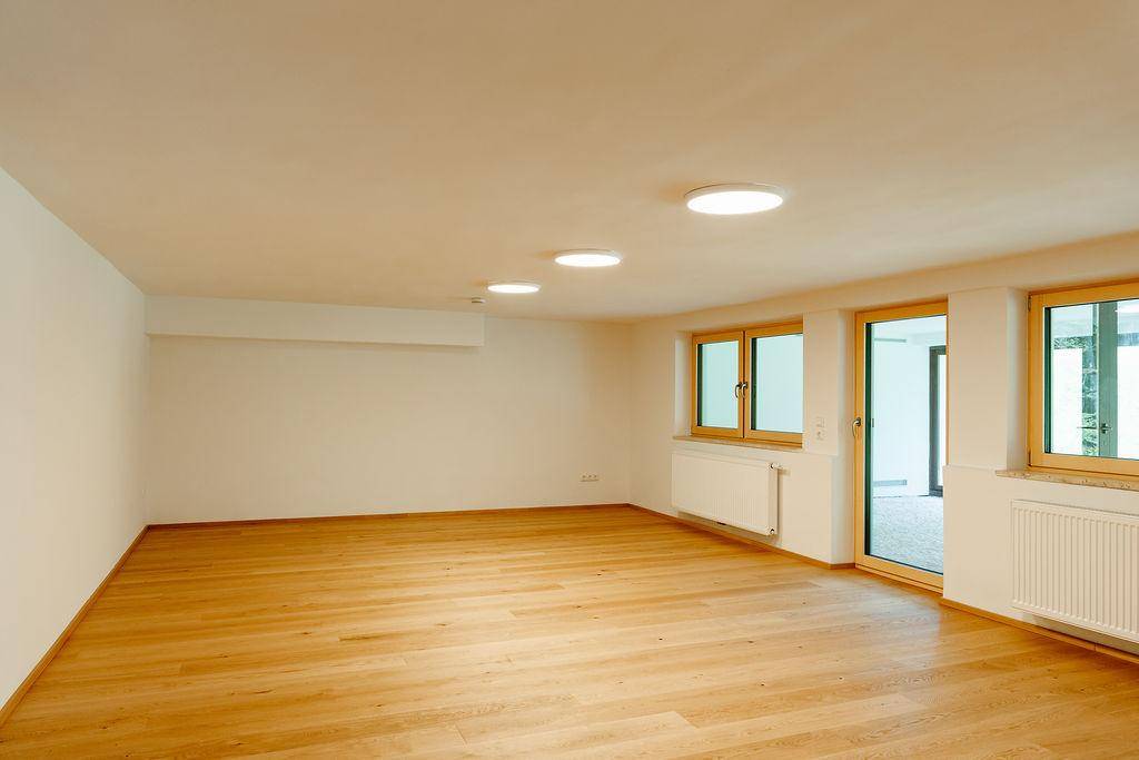 Geräumiger Wohnbereich mit Holzboden und großen Fenstern, die viel Licht hereinlassen.