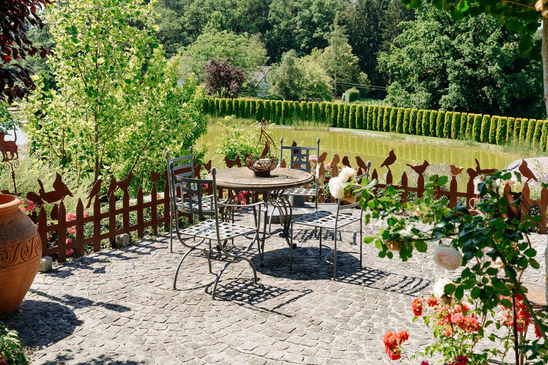 Gepflasterte Terrasse mit Gartenmöbeln und Blick auf den Teich, umgeben von einer dekorativen Metallzaun.
