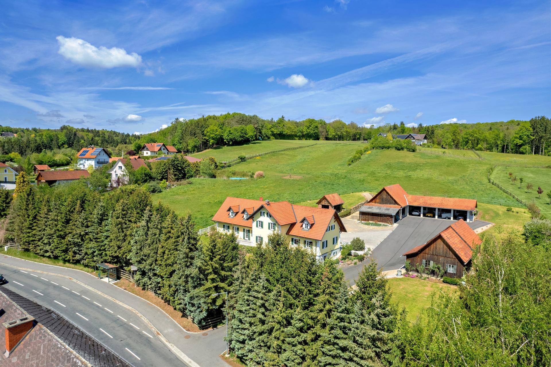 Ländliche Ansicht mit traditionellen Häusern, einer Straße und hohen Nadelbäumen vor grünen Hügeln.