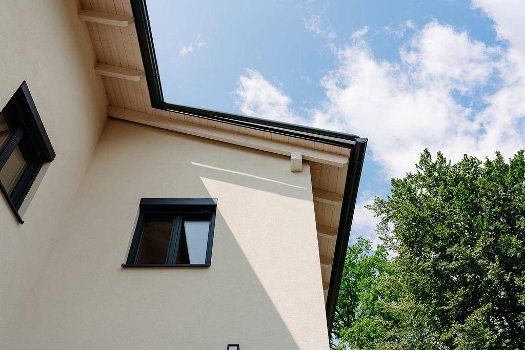 Nahaufnahme der Hausfassade mit Fenster, Rollladen und sichtbaren Dachbalken unter blauem Himmel.
