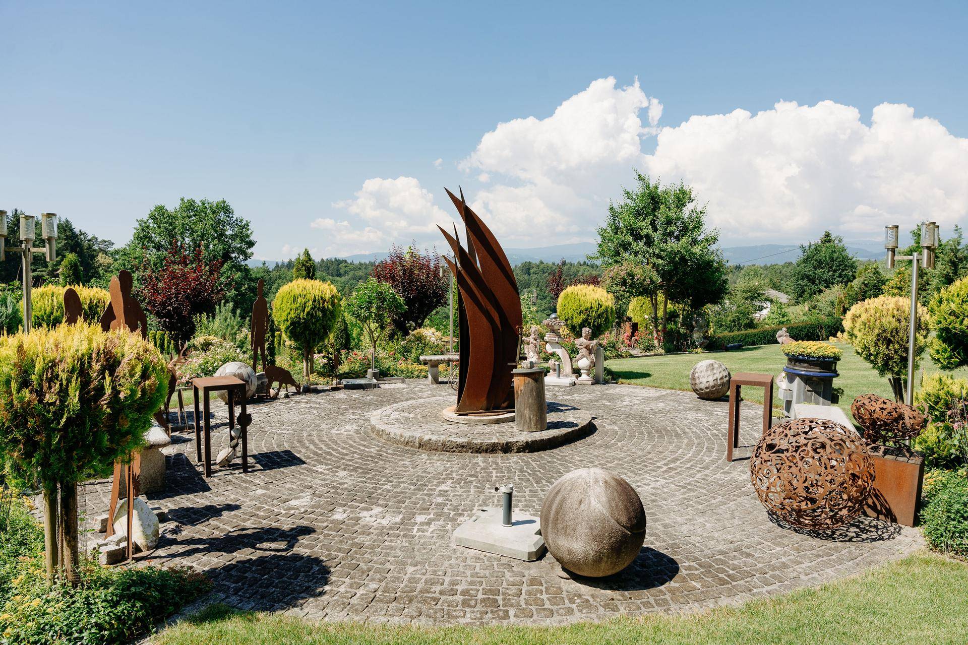 Künstlerisch gestalteter Gartenbereich mit modernen Skulpturen und gepflastertem Rundweg unter blauem Himmel.
