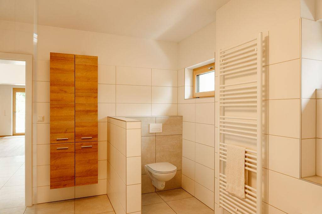 Modernes Badezimmer mit Wand-WC, Holzschrank und Handtuchheizkörper, hell gefliest.